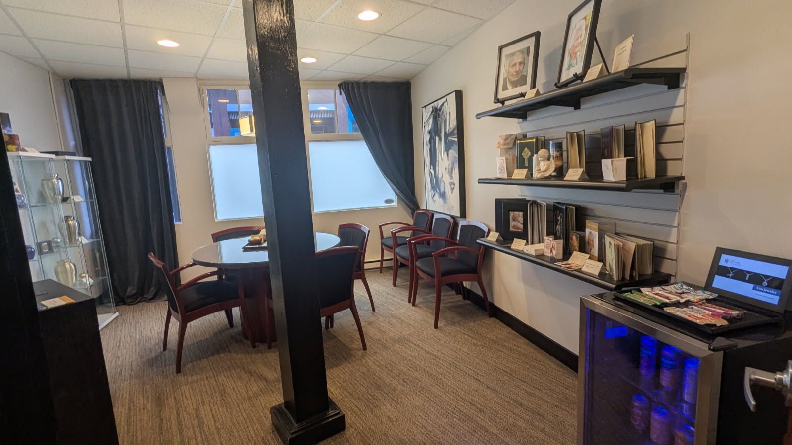A professional office interior with a round conference table, chairs, wall shelving with decor, and a small glass fridge.
