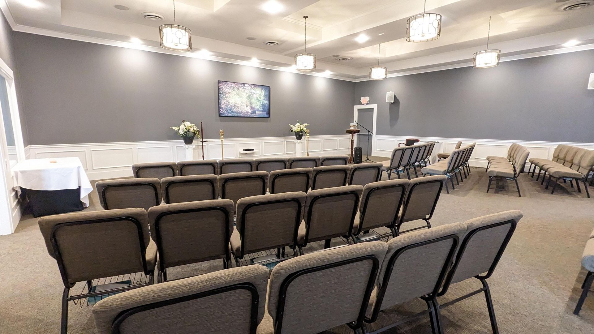 A community room or small hall with rows of gray chairs, a podium, and a wall-mounted television screen.
