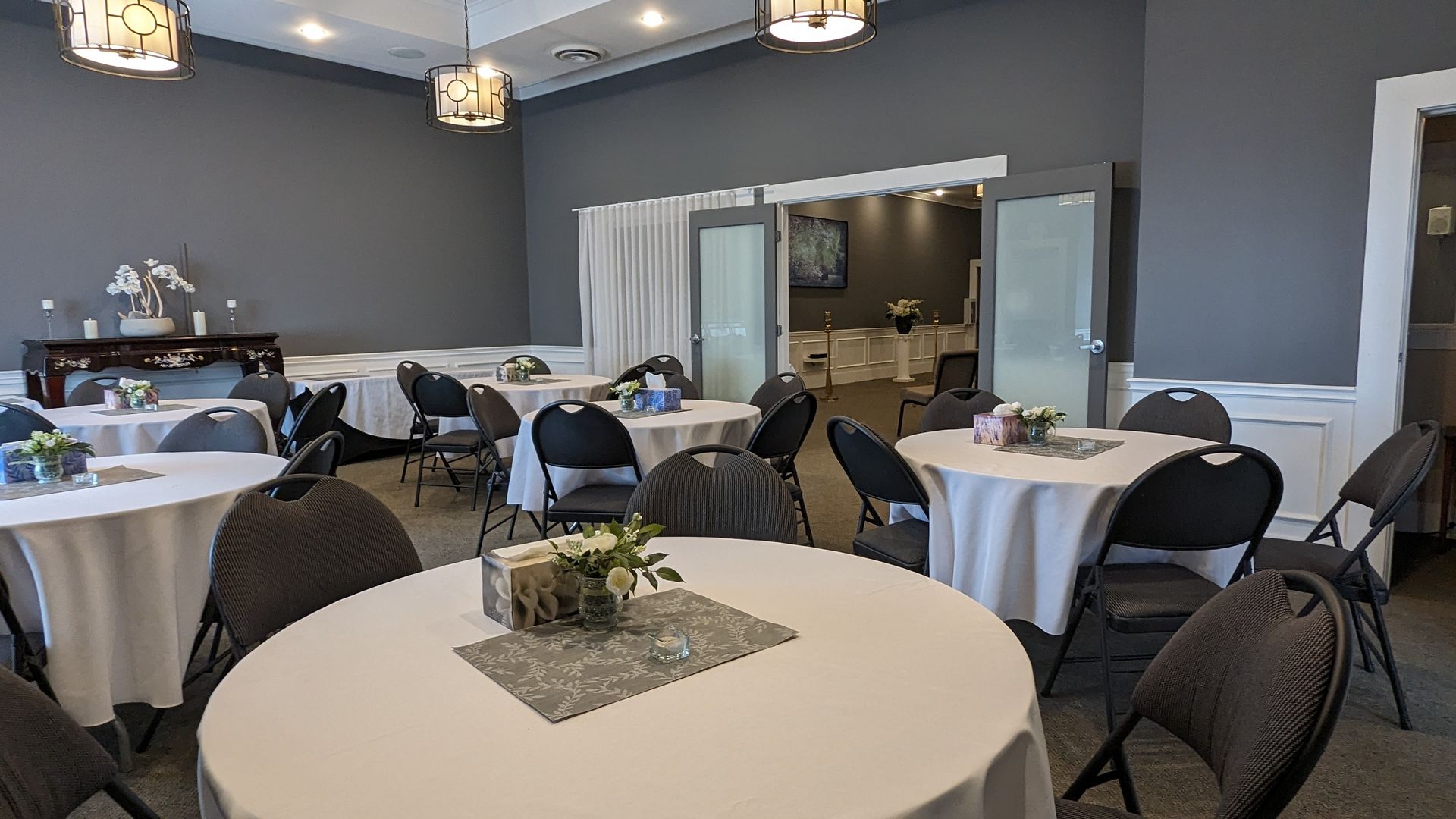 A banquet room with round tables, white tablecloths, dark chairs, gray walls, and glass-paneled double doors.
