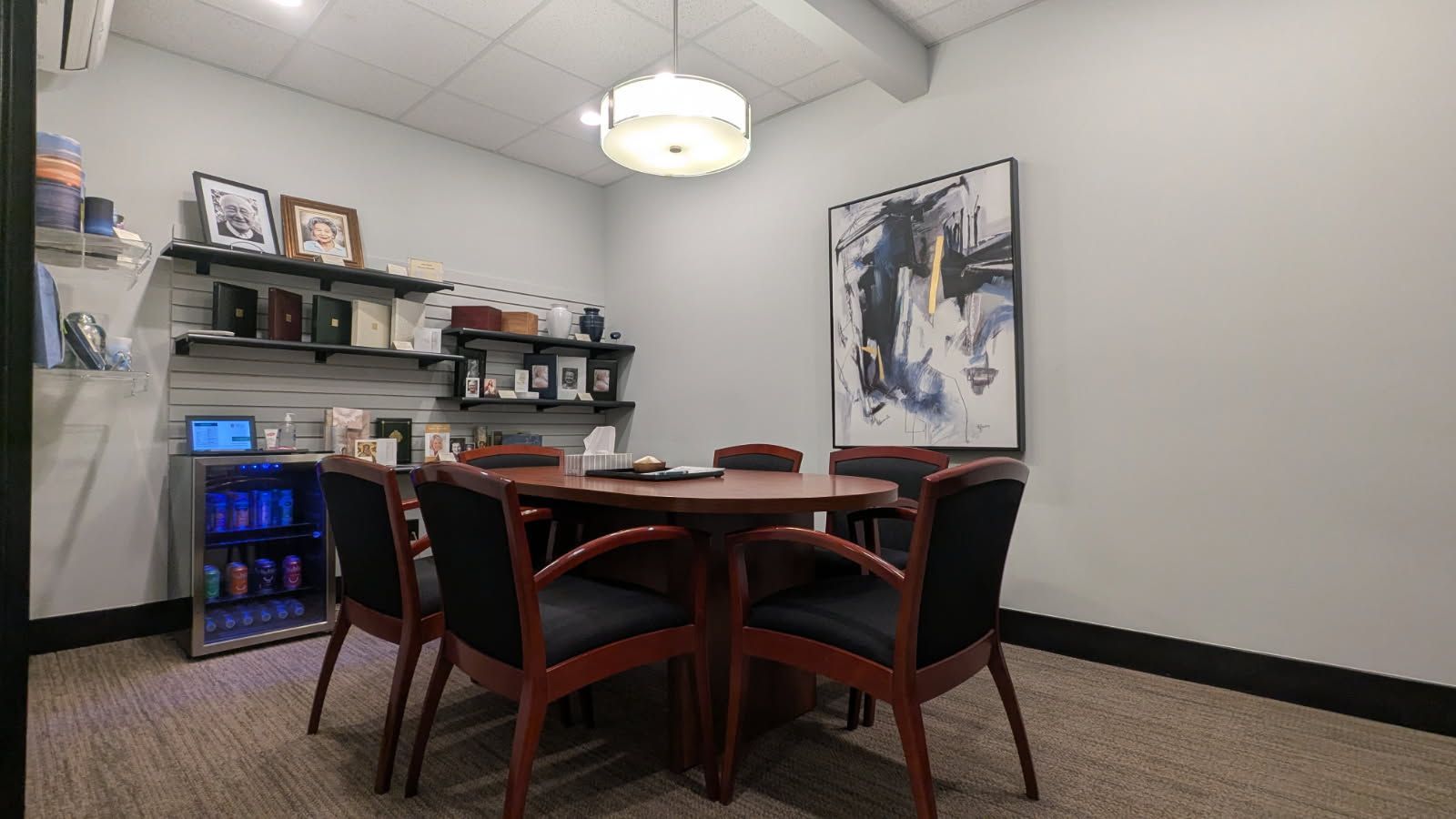 A small, modern meeting room with a wooden table, four chairs, shelves of decor, a beverage fridge, and wall art.