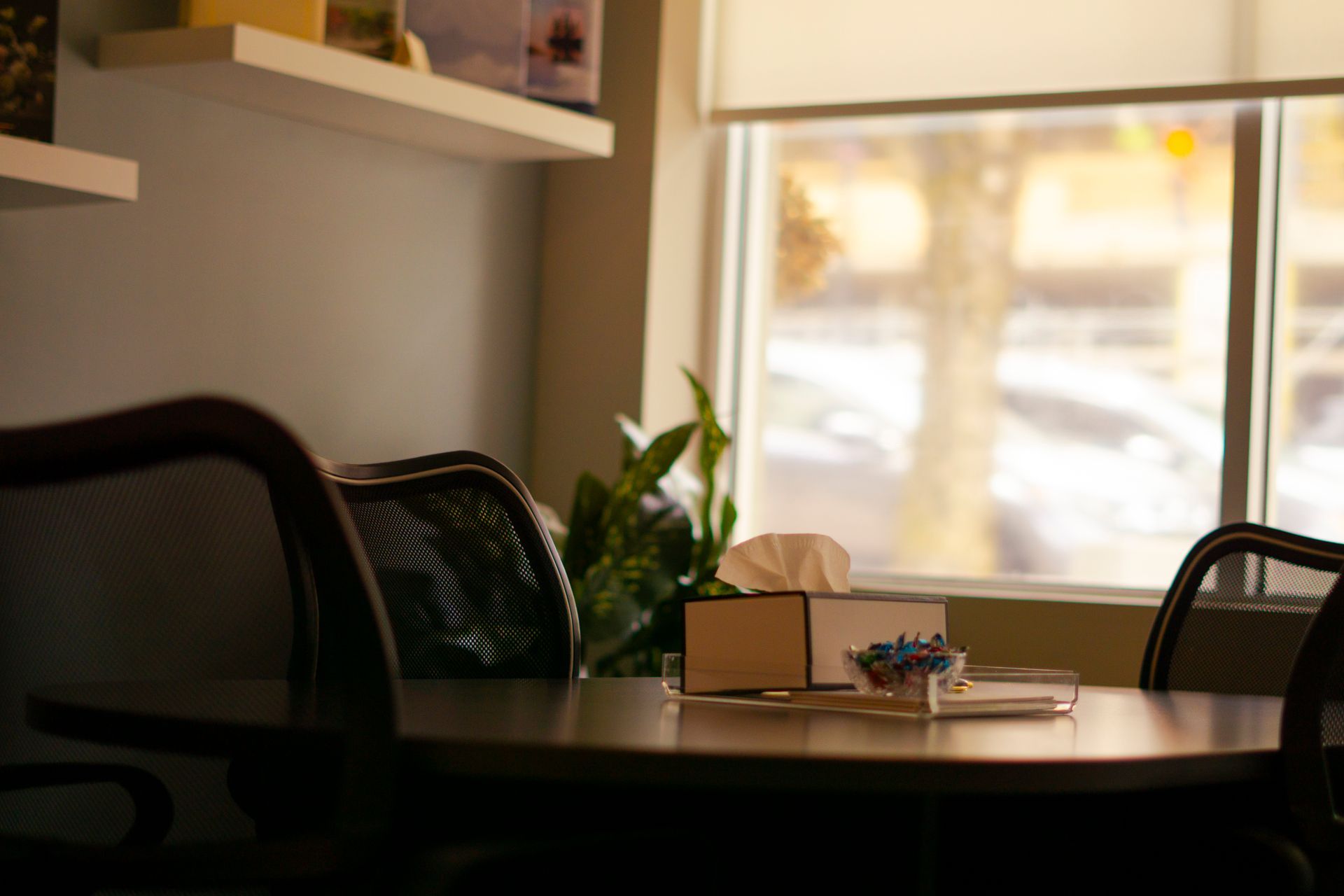 A table with a tissue box and small snack container sits in a room with a window view and floating shelves.