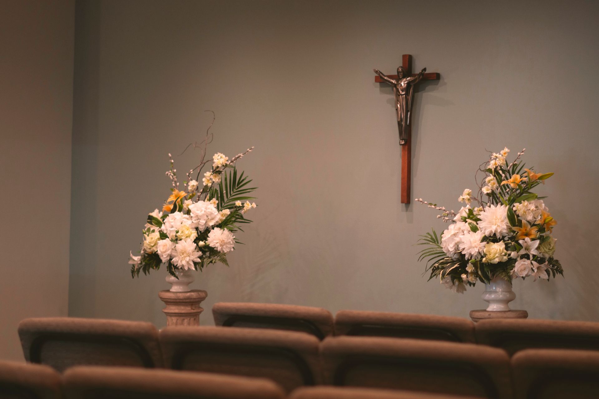A crucifix hangs on a gray wall behind two floral arrangements on stands, viewed from behind rows of church chairs.