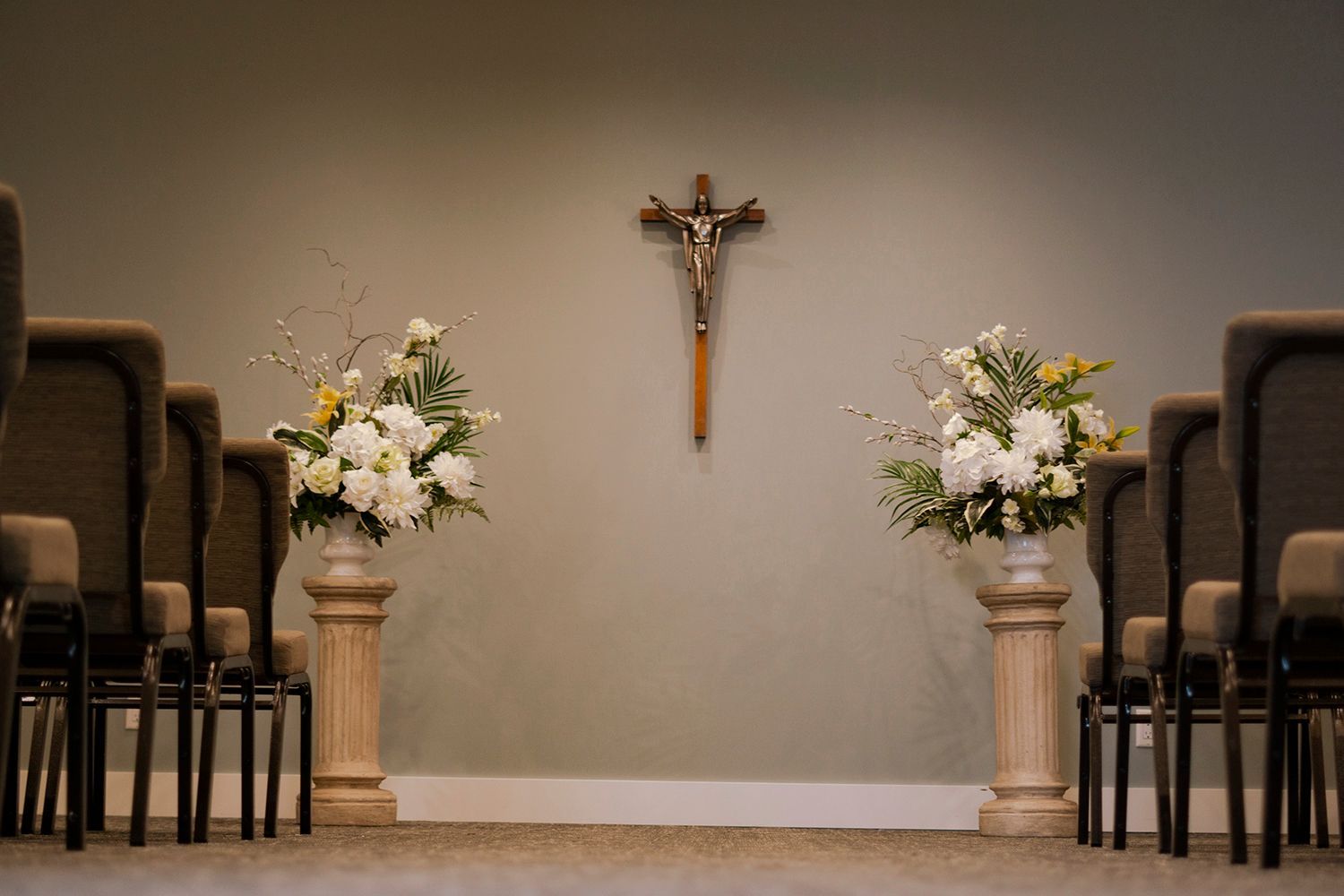 A crucifix on a plain wall centered between two floral arrangements on pedestals, flanked by rows of chairs in a room.