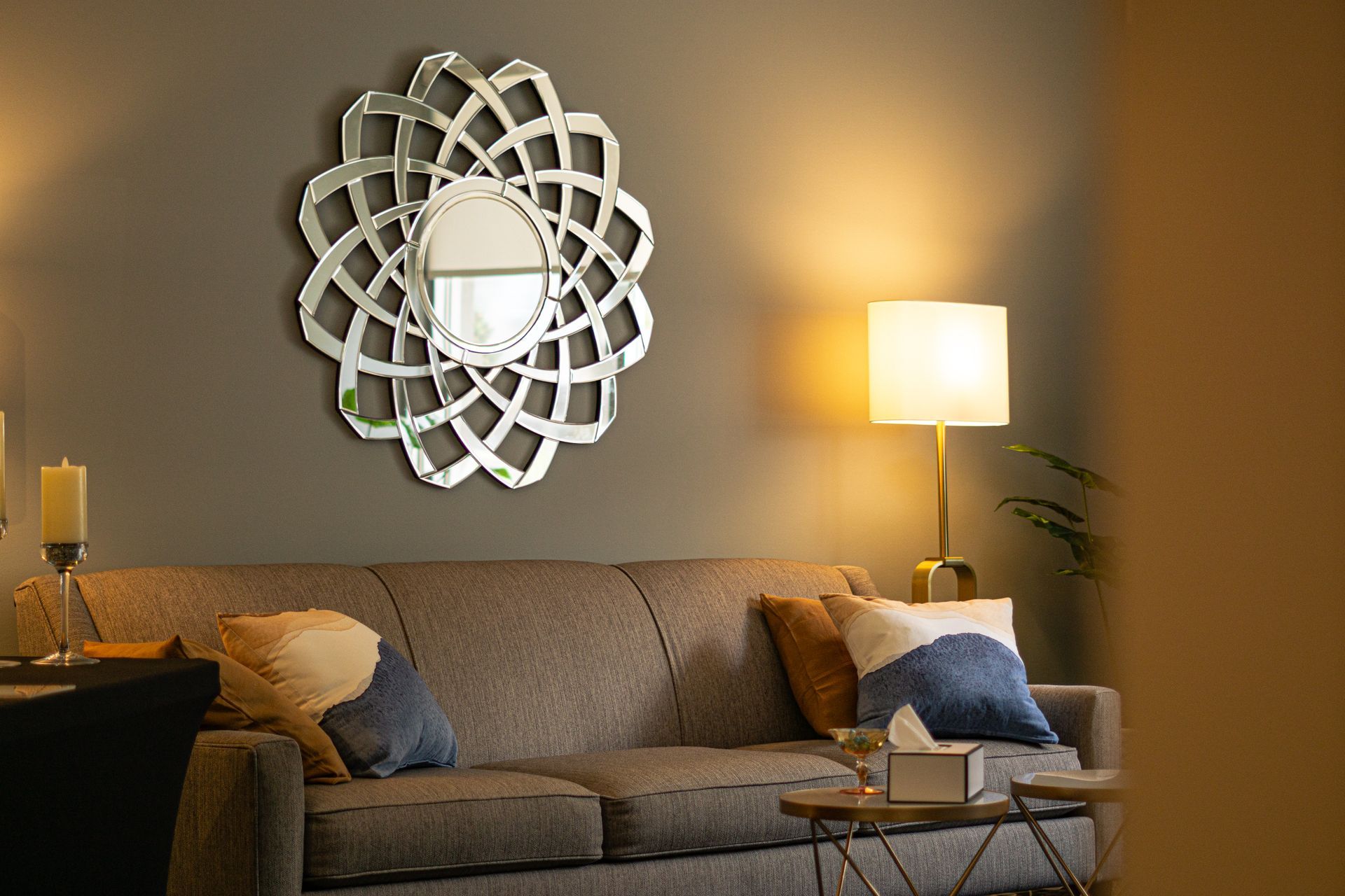 A grey sofa with colorful throw pillows sits below a decorative circular sunburst mirror on a muted grey wall.