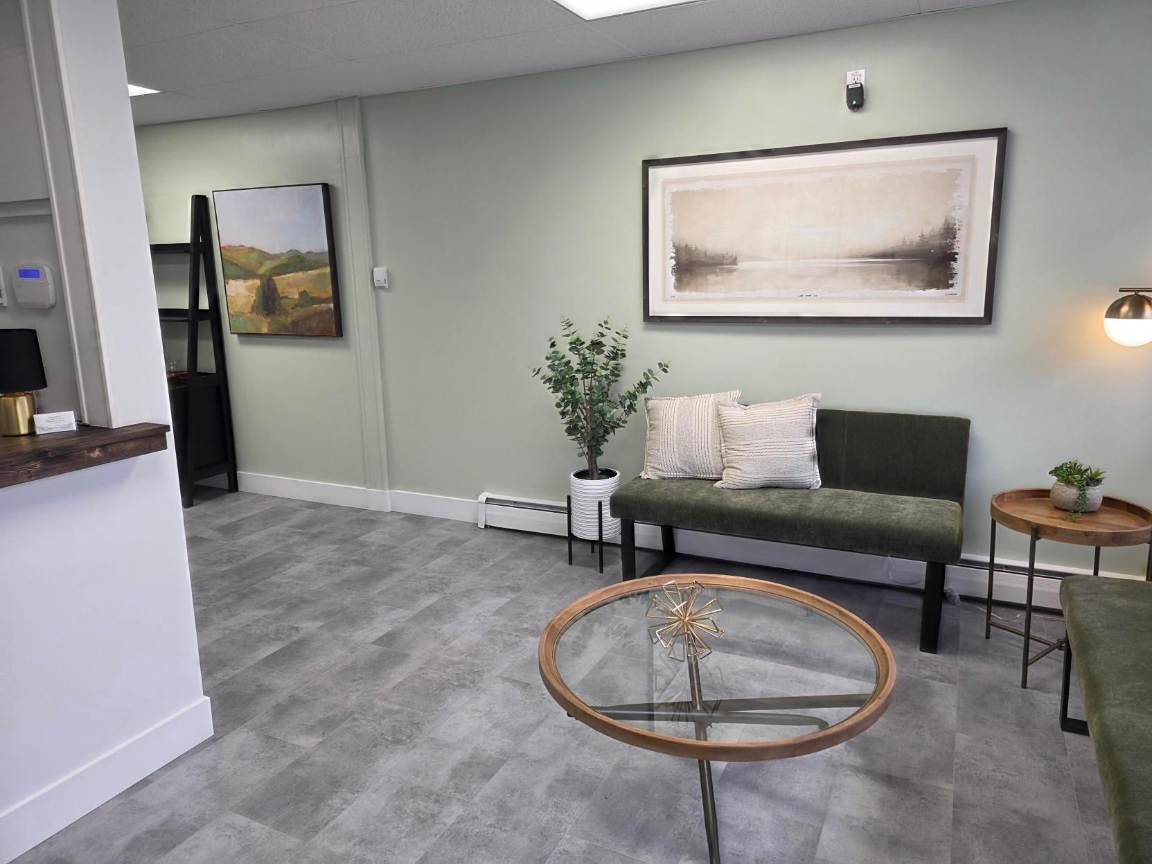 A waiting area featuring a green bench with pillows, a glass coffee table, wall art, and a plant on a light green wall.