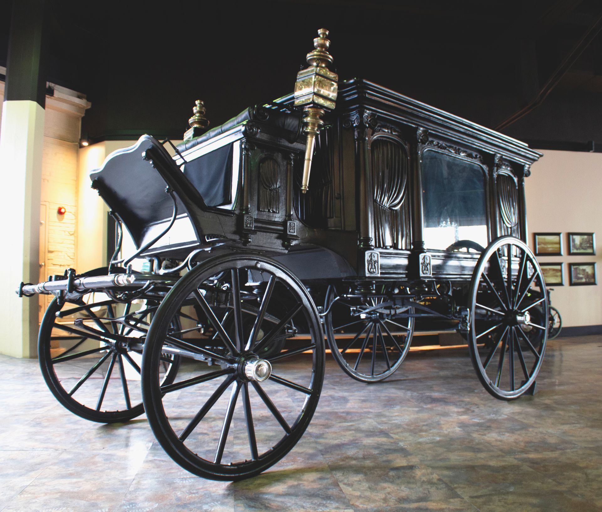 An ornate, black horse-drawn funeral carriage with large wooden-spoked wheels, displayed in a museum gallery.