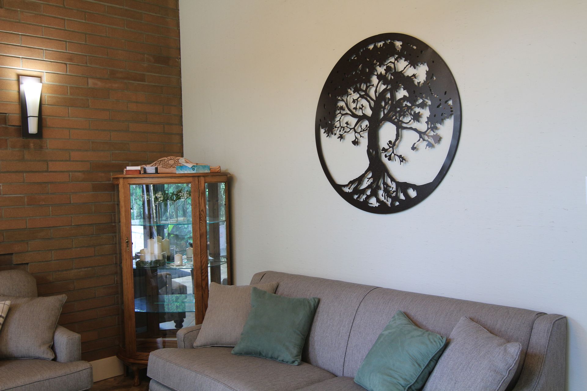 A living room with a gray sofa, a wood display cabinet, and a black circular metal tree wall sculpture.