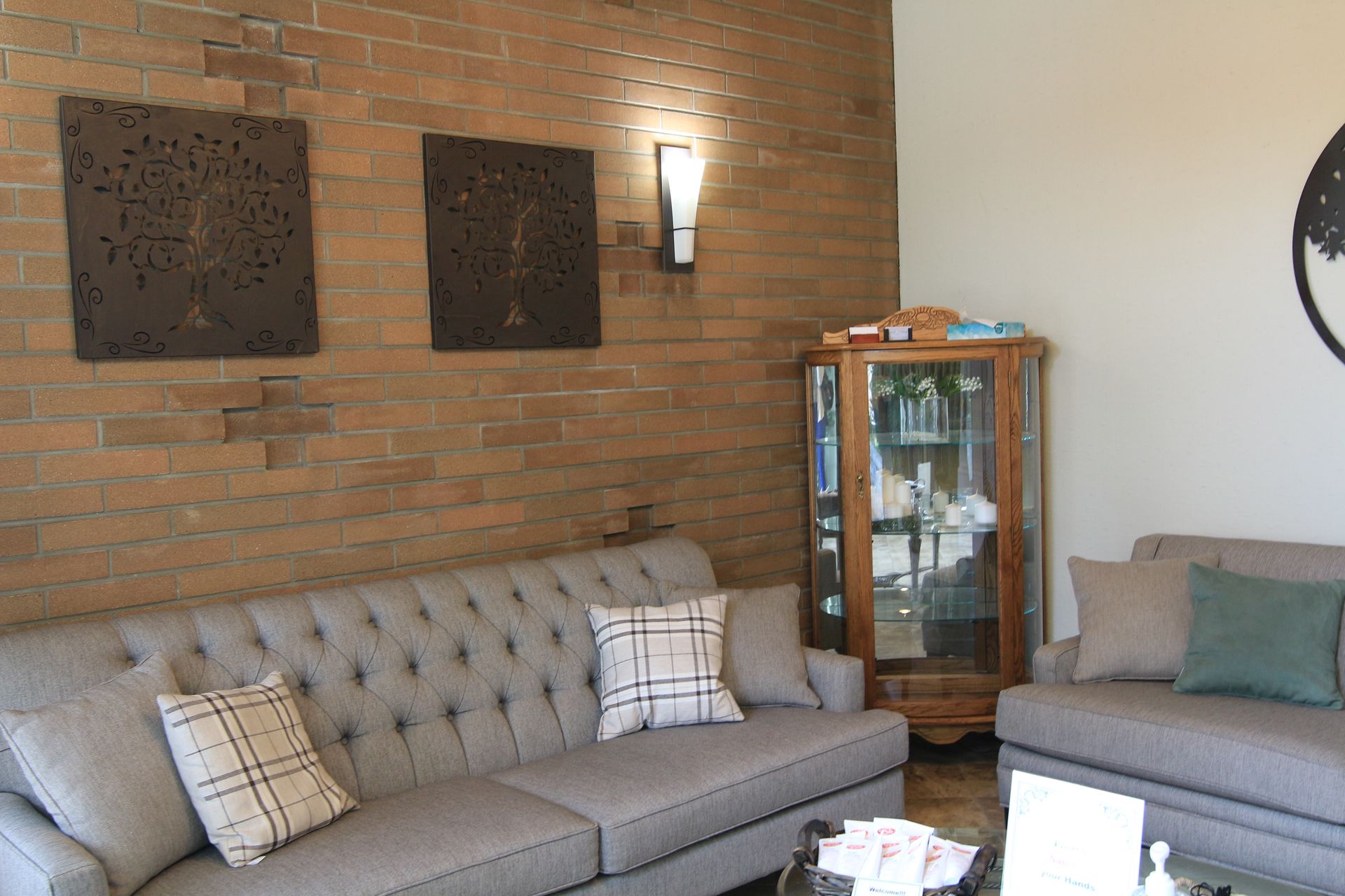 A living room with a brick wall, two gray couches, a corner display cabinet, and tree-themed wall art.