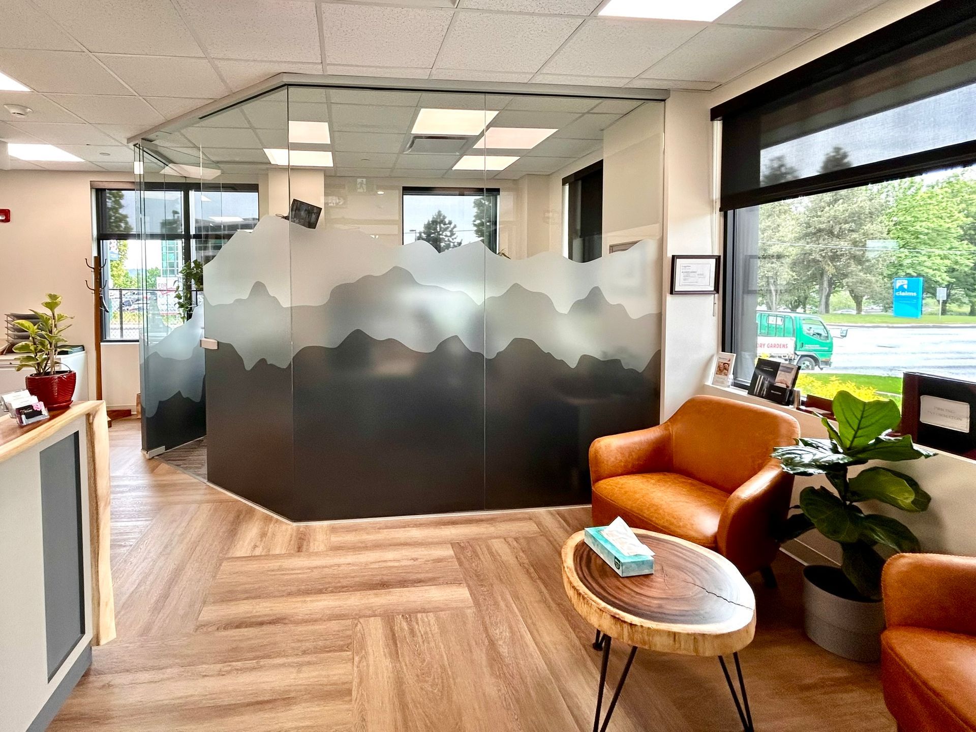 A modern waiting area with tan armchairs, a wood-slice coffee table, and a glass partition featuring a mountain design.