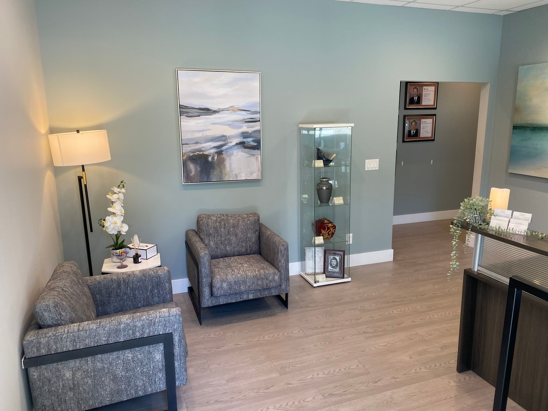 A waiting area with light blue walls, two patterned grey chairs, a floor lamp, a display cabinet, and abstract art.