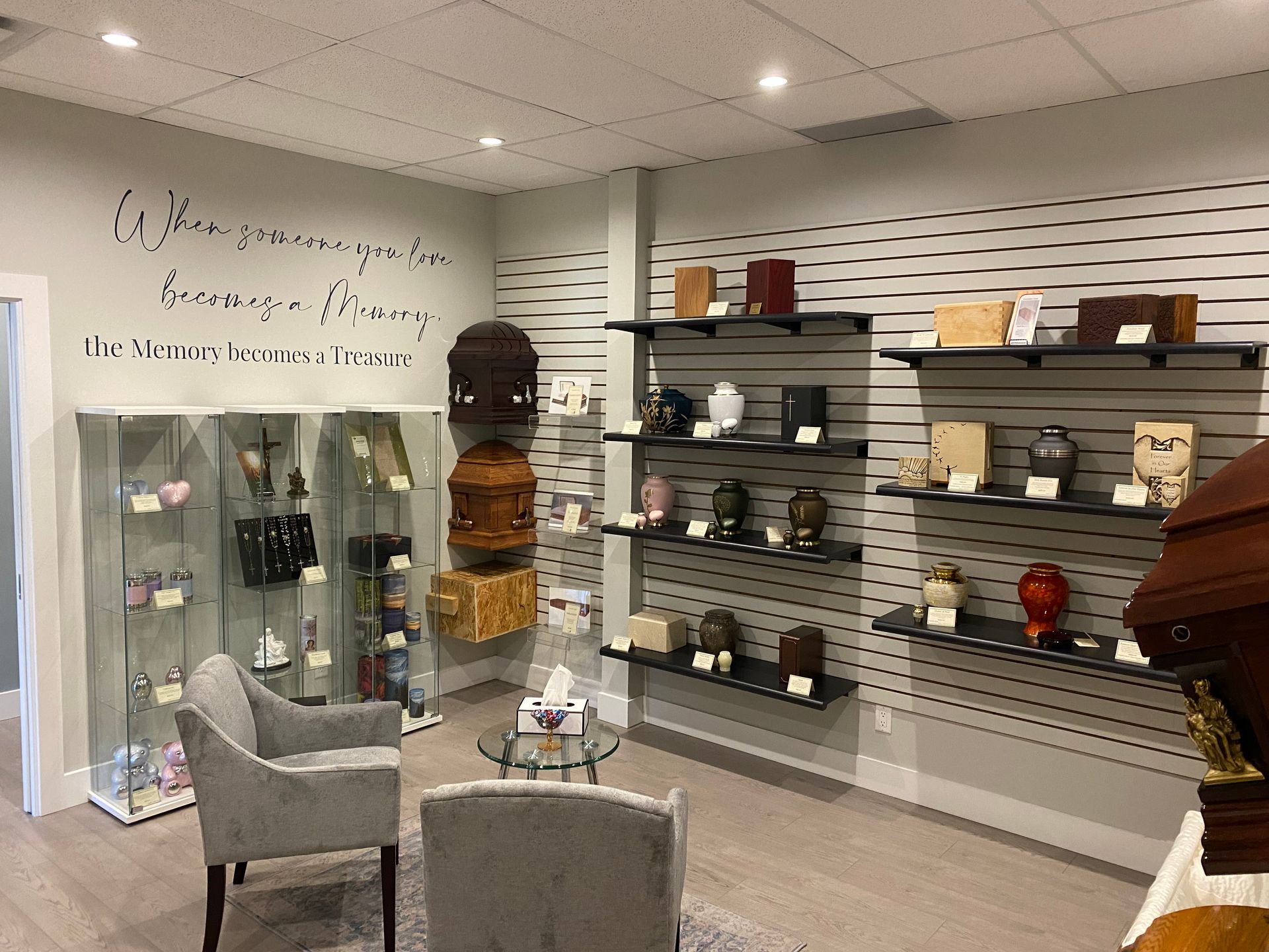 A funeral home showroom featuring urns on wall-mounted shelves, display cases, and two gray armchairs for consultation.