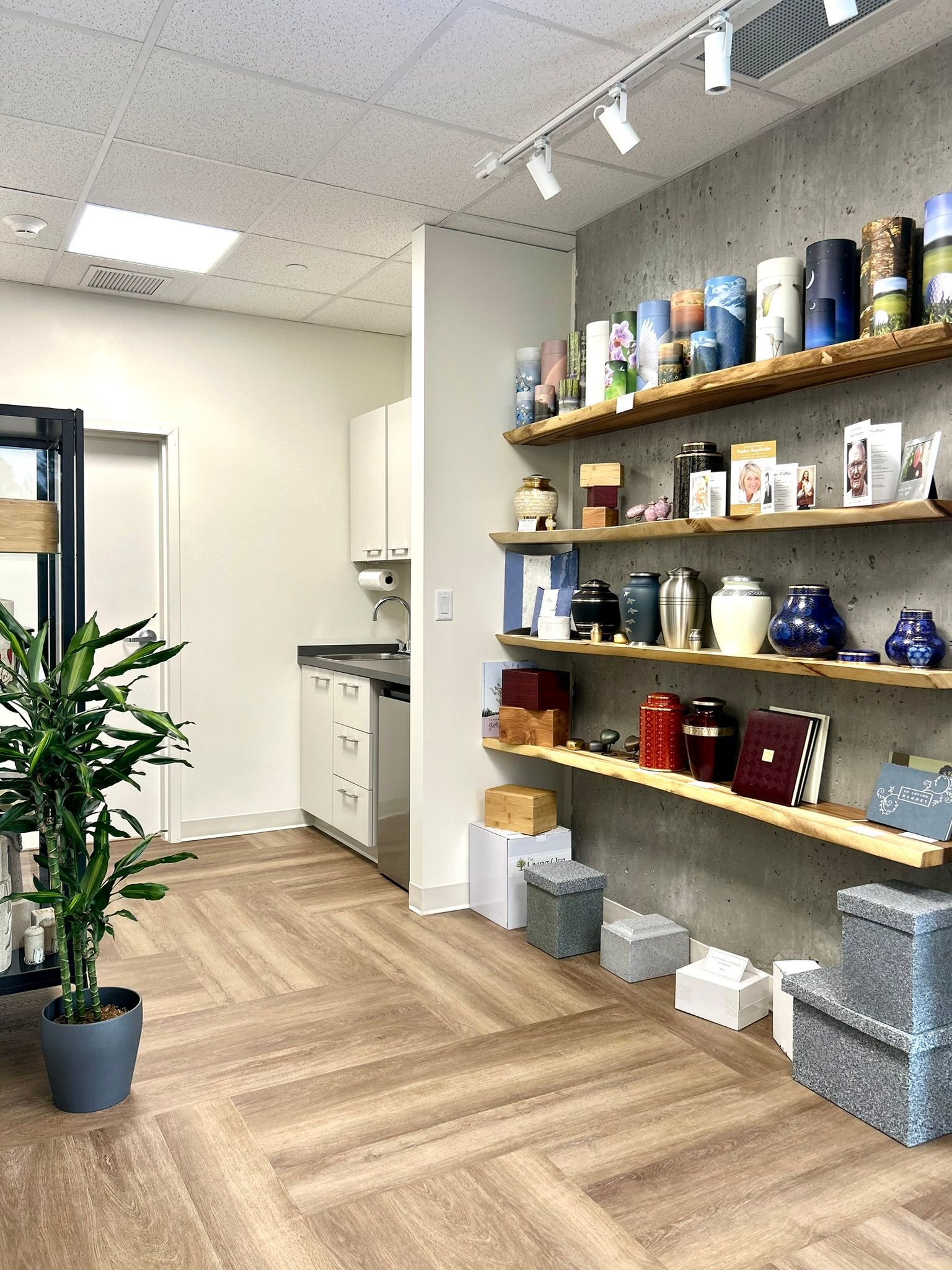 A display of various urns and memorial items on wooden shelves against a concrete wall inside a well-lit showroom.