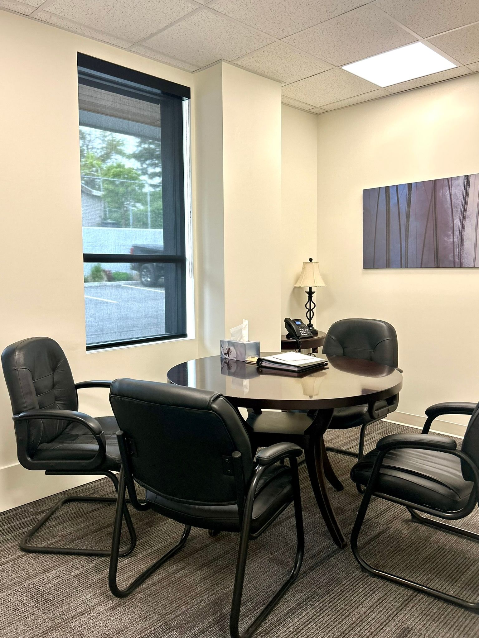 A small meeting room featuring a round table, four black office chairs, a window, a table lamp, and wall art.