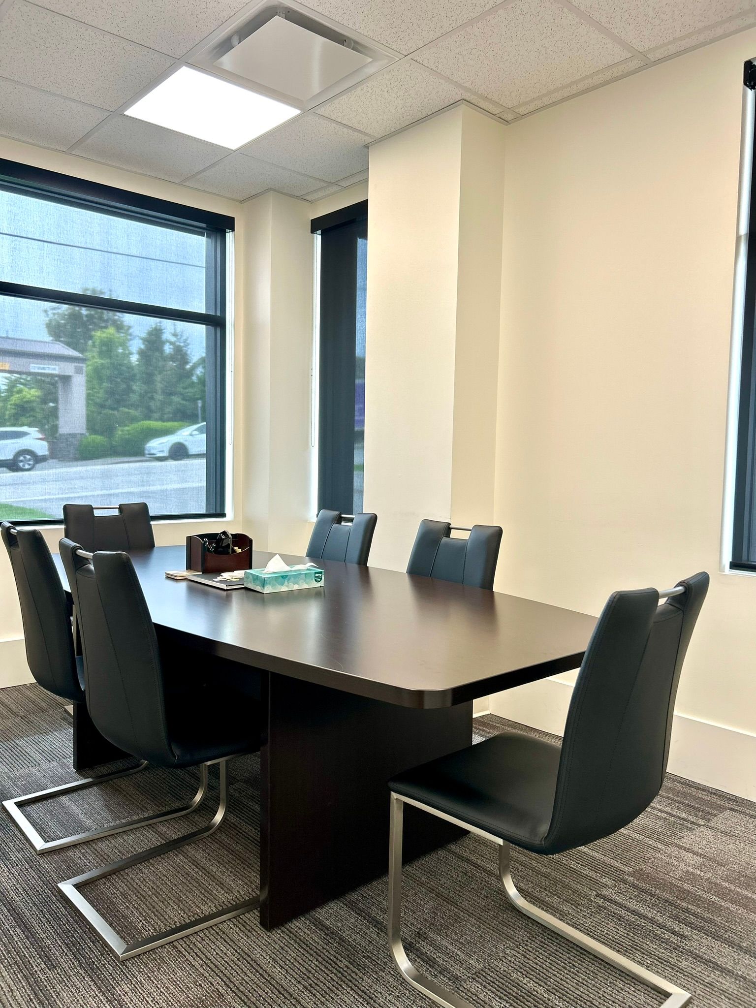 A modern, empty conference room with a long brown table, six black chairs, beige walls, and large windows.