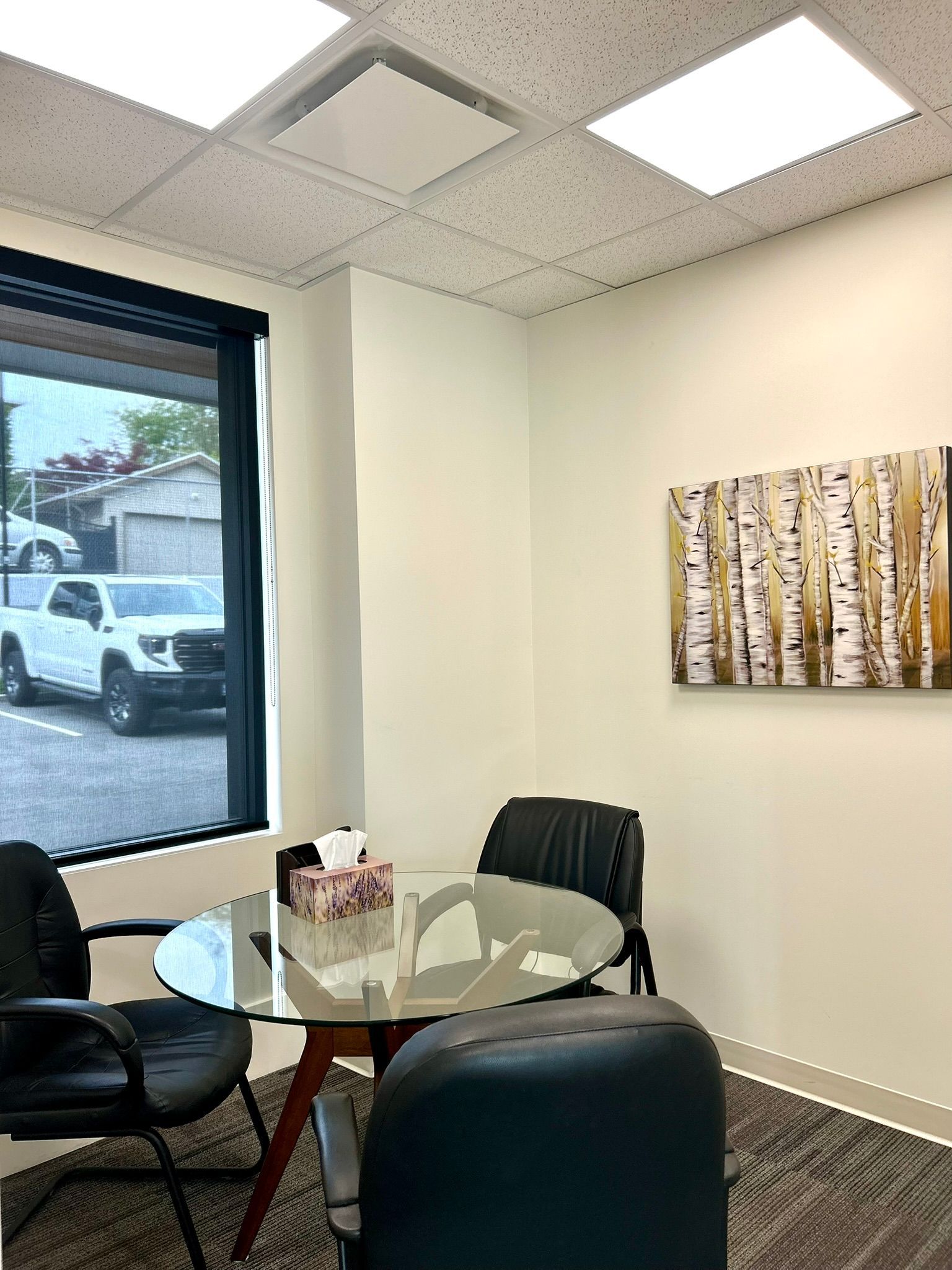 A small office with a glass round table, black chairs, a window view of a parking lot, and birch tree wall art.