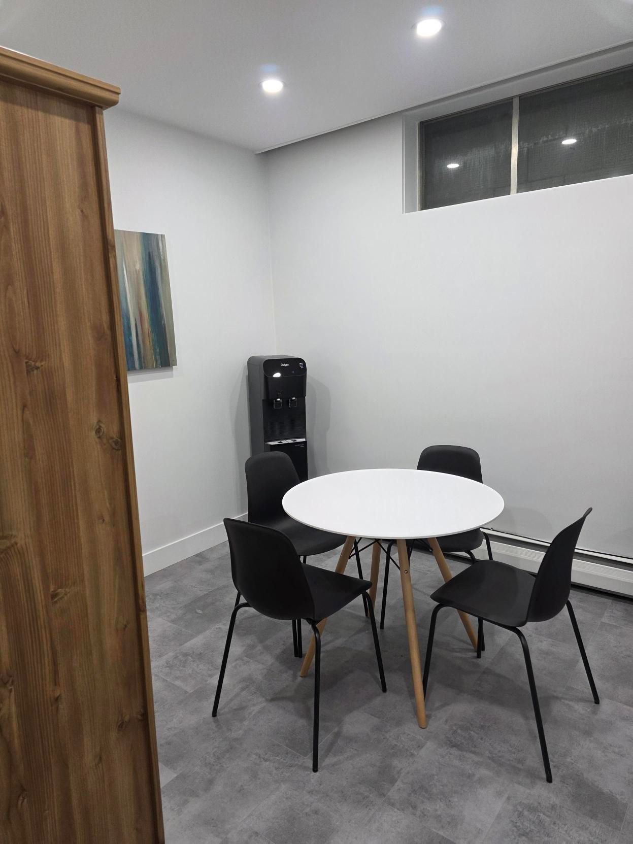 A small, modern breakroom with a round white table, four black chairs, a water cooler, and art on a light gray wall.