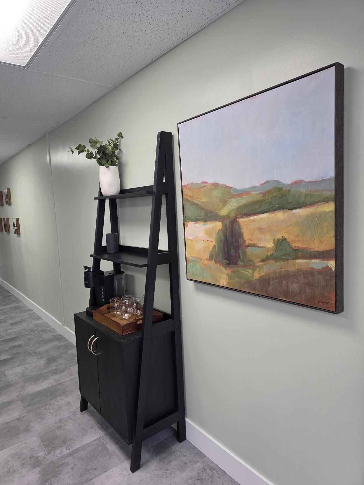 A black ladder shelf with a coffee station stands against a pale green wall next to a large landscape painting.