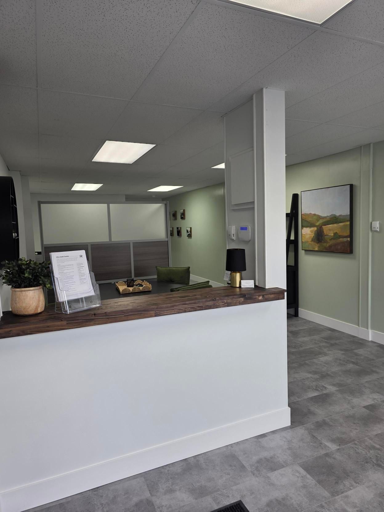 A modern office reception area with a white front desk, gray tile floors, pale green walls, and a landscape painting.