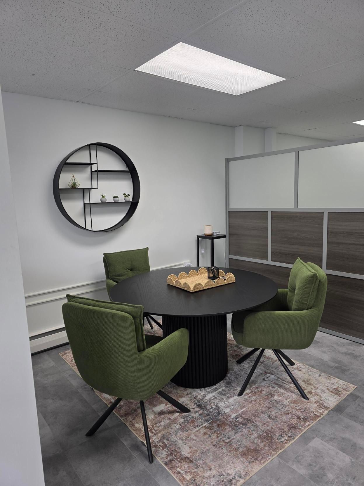 A round black table with three green chairs and a tray sits on a patterned rug in a modern office with a circular shelf.