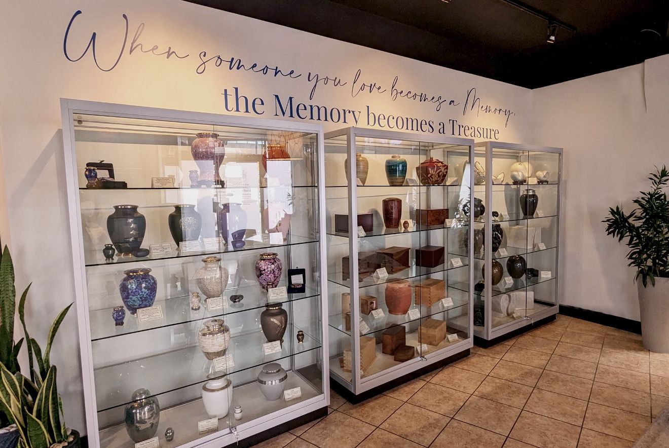 Three glass display cases filled with decorative urns stand under a wall quote about memories in a funeral home.