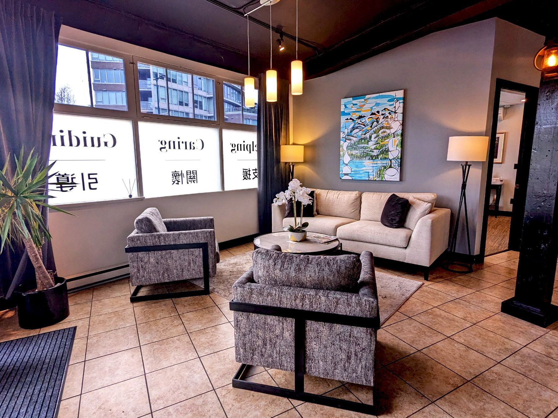 A modern, neutral-toned lobby with two patterned armchairs facing a light beige sofa, large windows, and pendant lights.