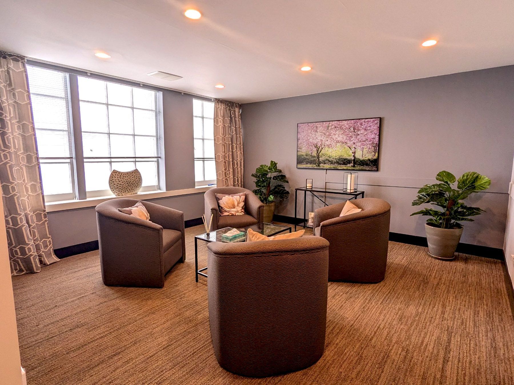 A seating area with four brown armchairs arranged around a glass coffee table in a room with grey walls and large windows.