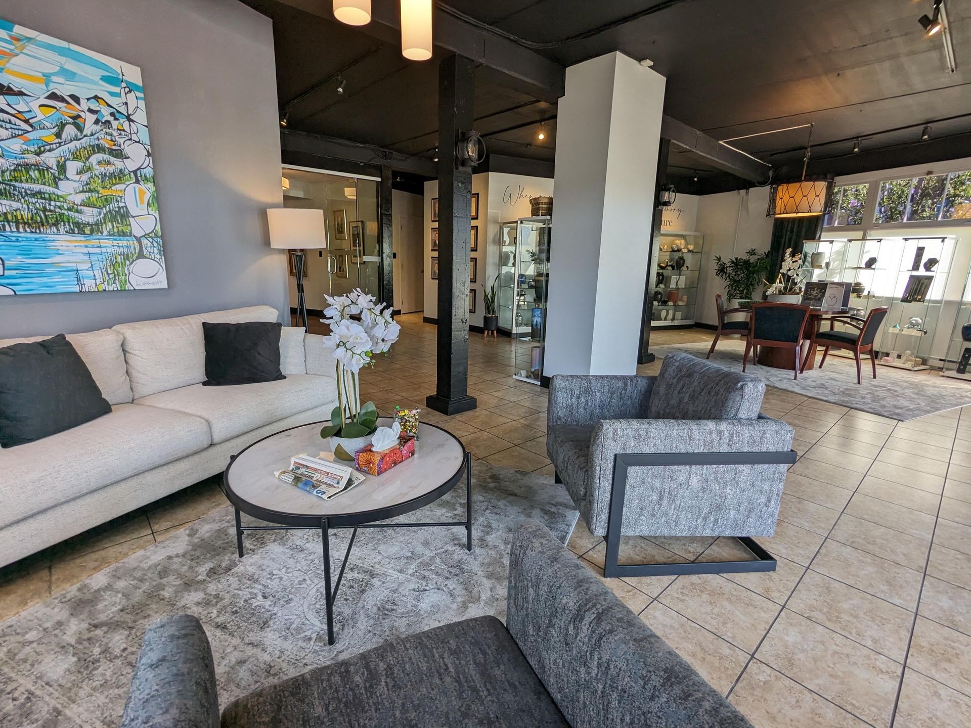 A modern office waiting area features a beige sofa, two patterned gray armchairs, and a round coffee table on a rug.