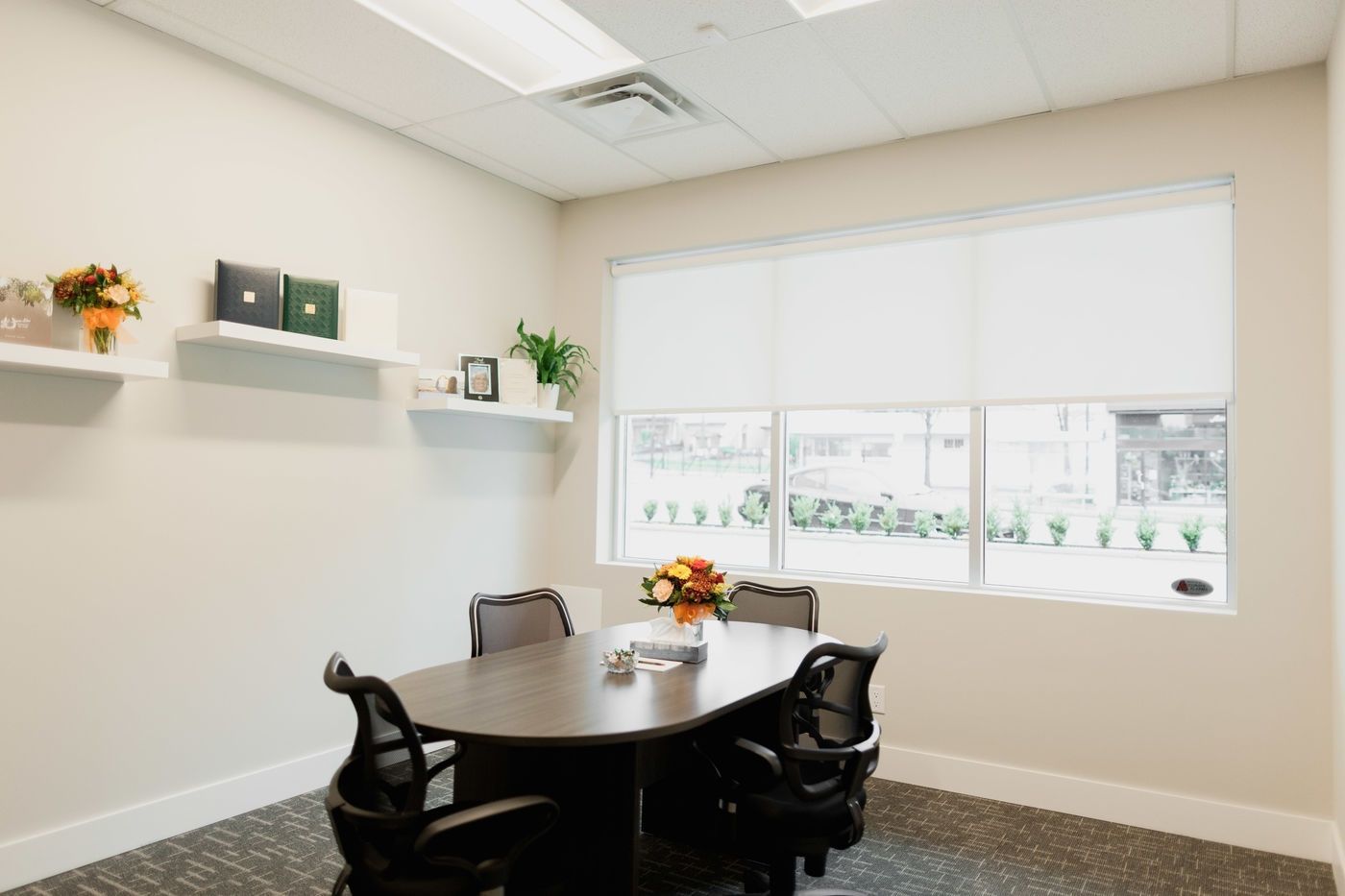 A small, modern meeting room with a dark oval table, four ergonomic black chairs, shelves with decor, and a large window.