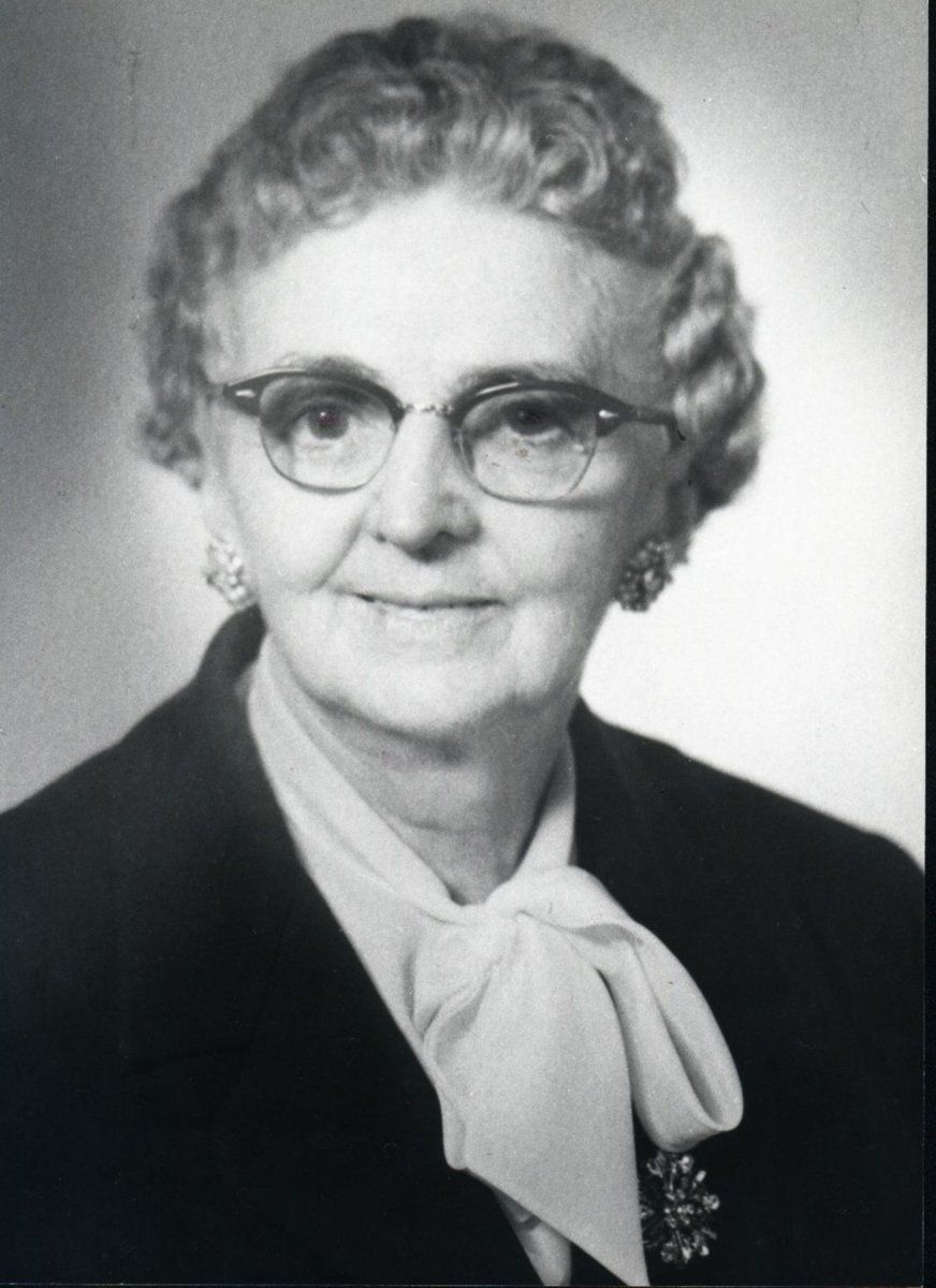 A portrait of an individual wearing glasses, earrings, a dark jacket, and a light-colored necktie blouse.