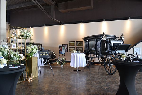 A funeral home showroom featuring a black horse-drawn hearse, floral arrangements, and pedestal tables on a stone floor.