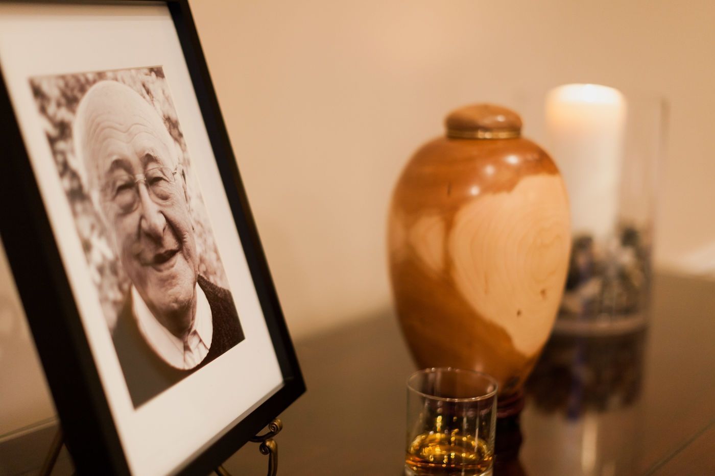 A memorial display with a framed portrait of an elderly person, a wooden urn, a glass of amber liquid, and a lit candle.