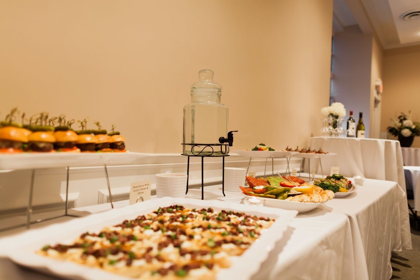 A buffet table with a glass water dispenser, sliders, flatbread, and various appetizers in an indoor setting.