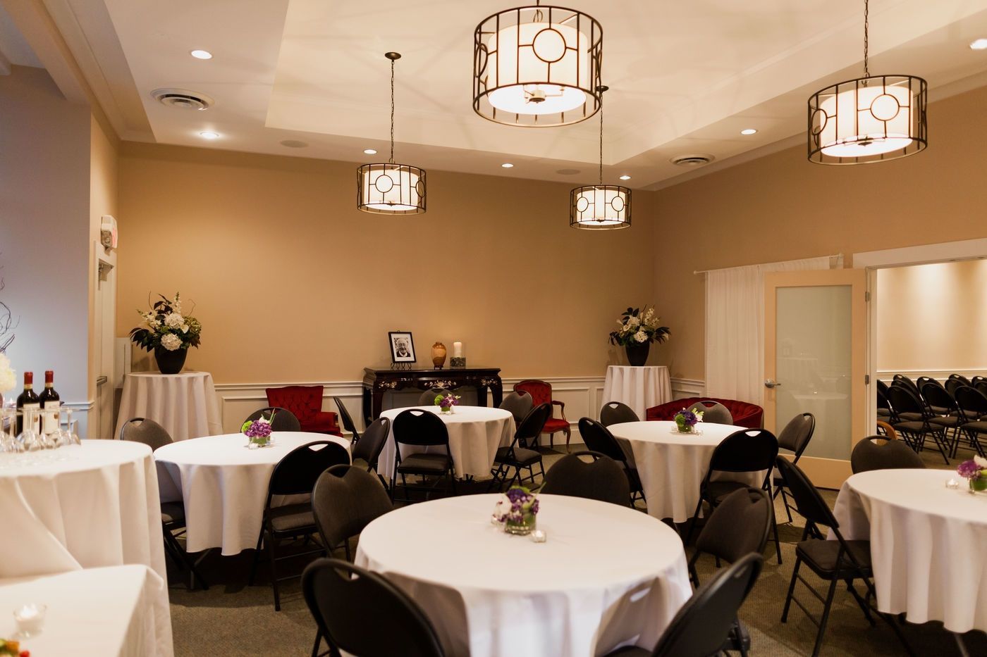 A banquet room with round tables, white tablecloths, and black chairs beneath decorative hanging pendant lights.