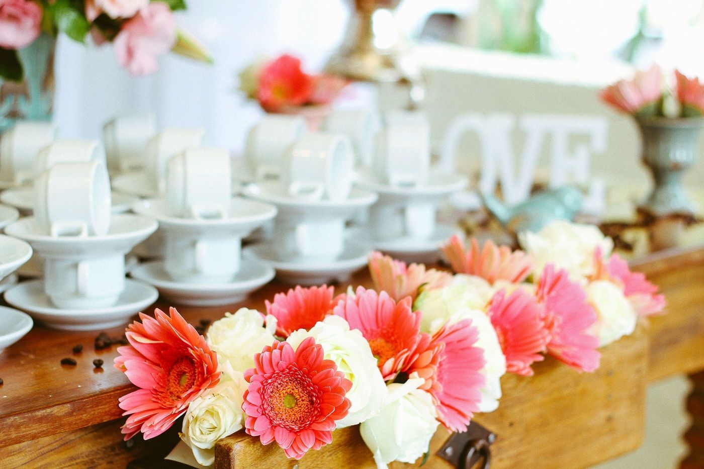 A wooden table holds stacks of white teacups and a vibrant garland of coral and white flowers with a