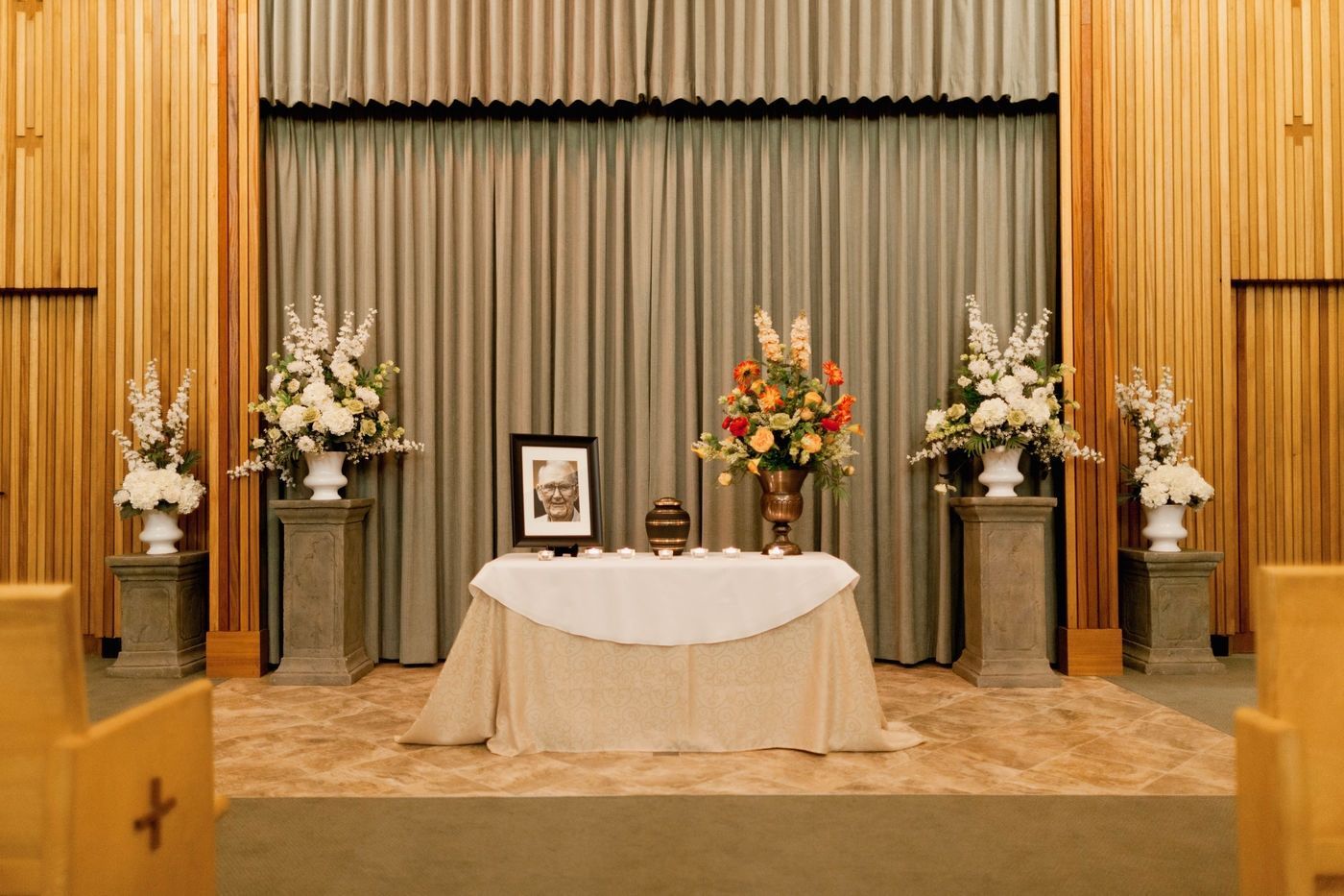 A memorial display with a framed photo, an urn, and floral arrangements on pedestals in front of a grey curtained wall.