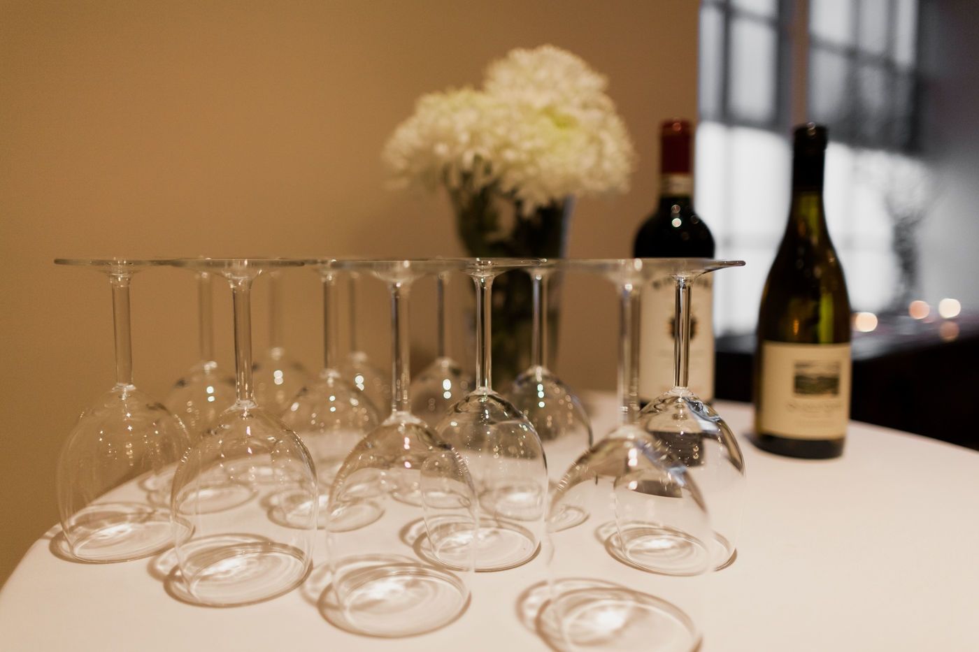 Several wine glasses arranged upside down on a white tablecloth, with a vase of white flowers and two wine bottles behind.