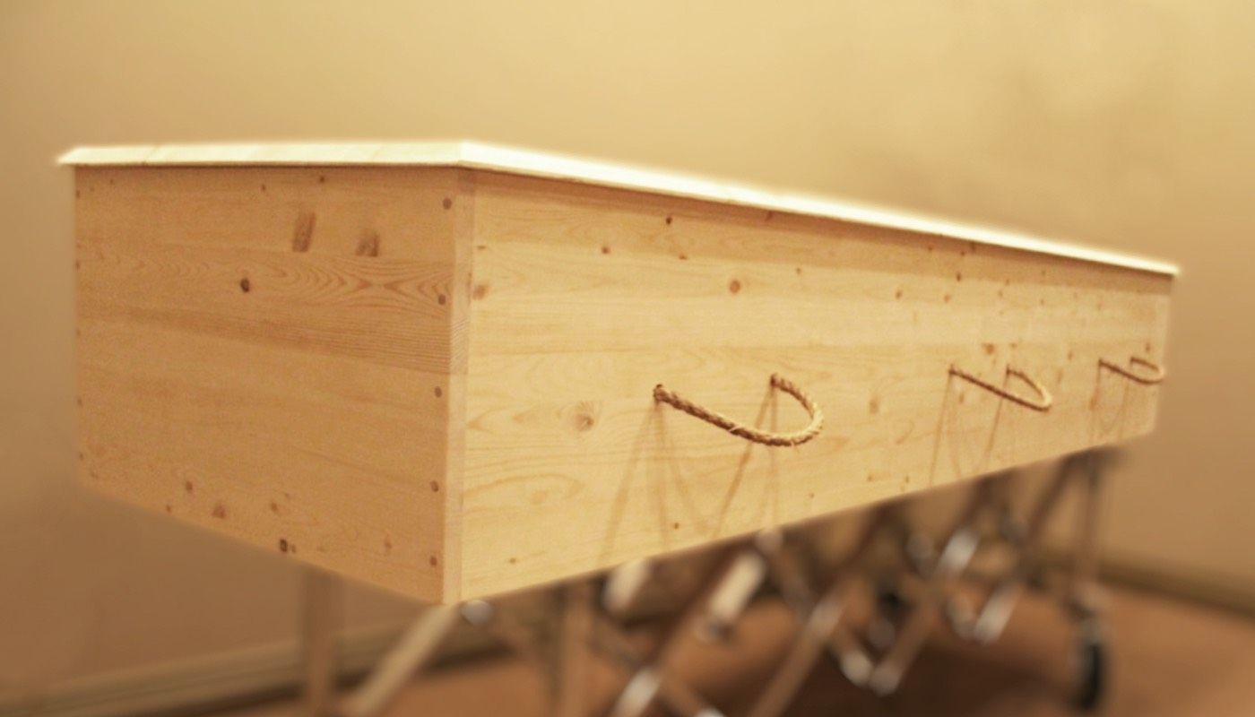 A simple, light-wood rectangular casket with three rope handles, resting on a folding metal stand indoors.