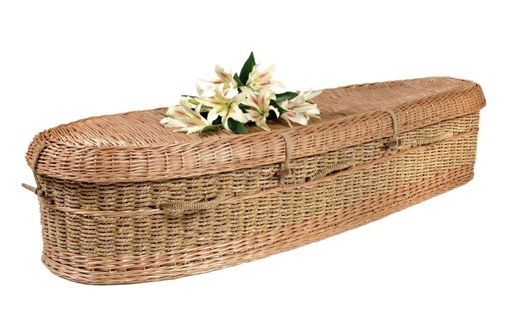 A tan woven wicker casket topped with a small bouquet of white lilies against a white background.