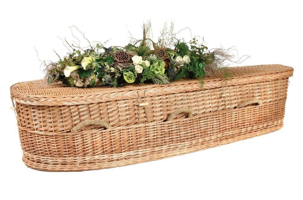 A woven wicker casket adorned with a floral spray of greenery and white flowers against a white background.