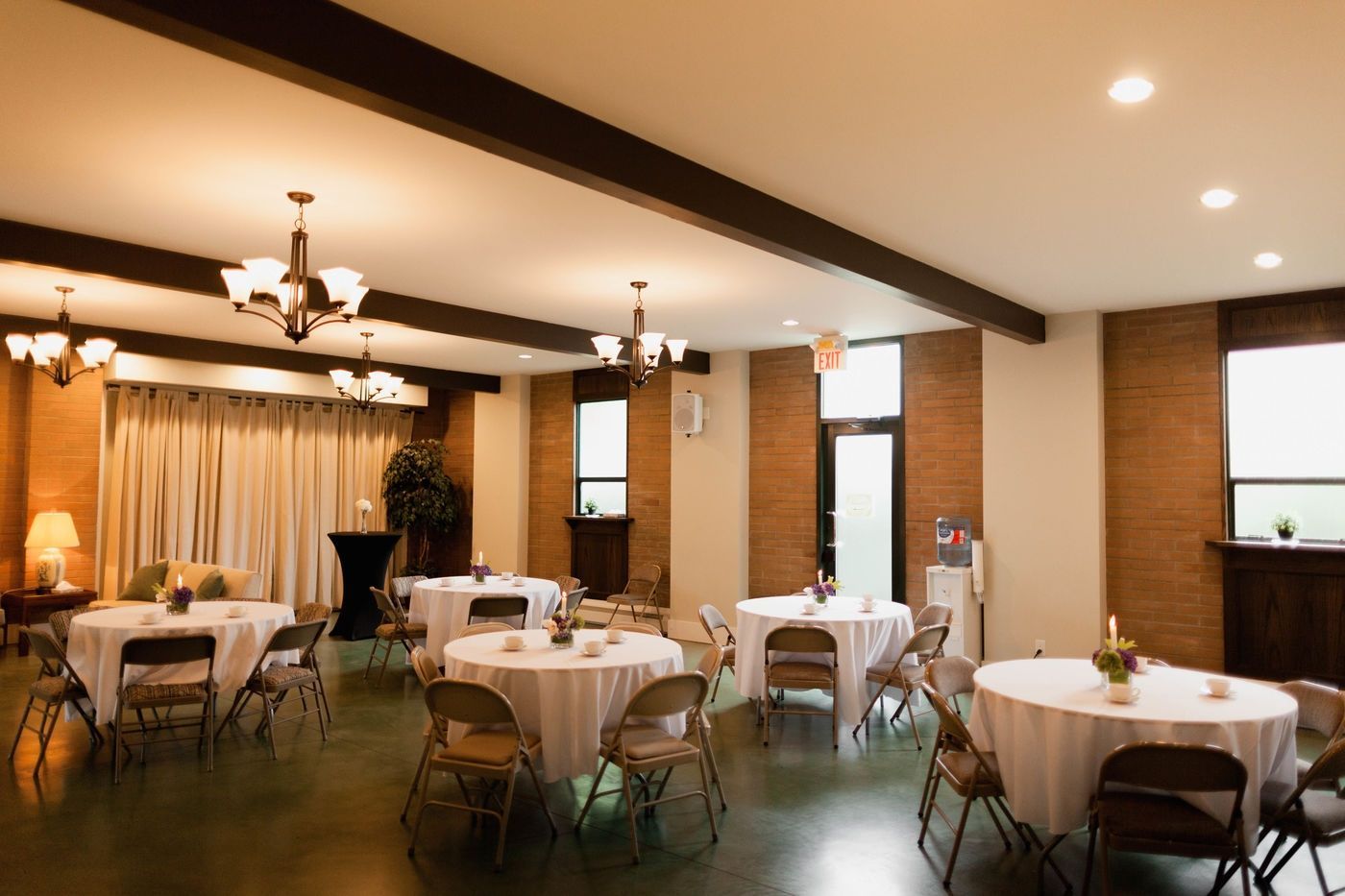 A banquet hall with several round tables draped in white tablecloths, brown chairs, chandeliers, and brick walls.