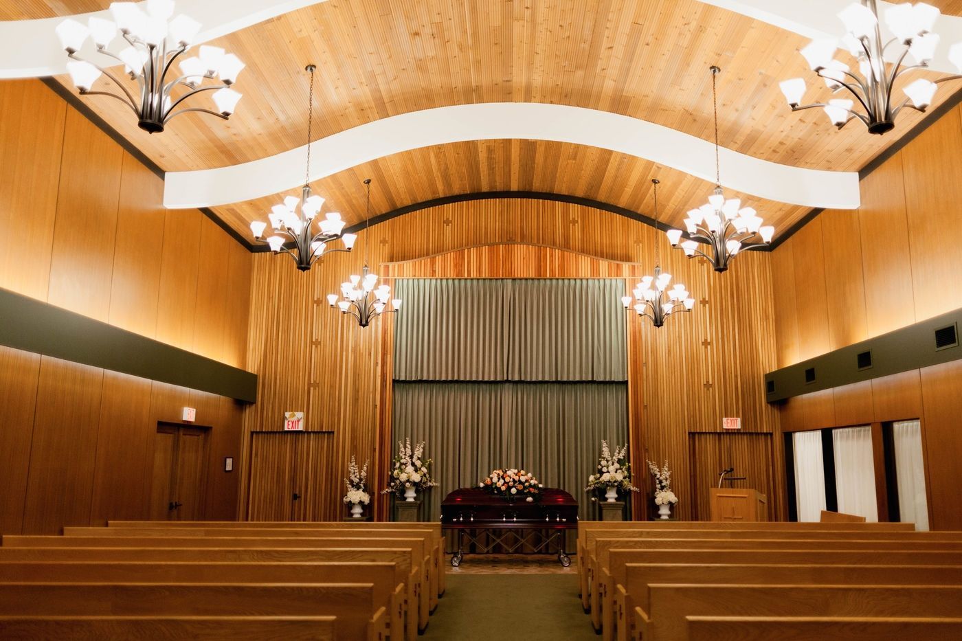 A funeral home chapel with wooden walls, rows of pews facing a central casket draped with flowers on a stage.