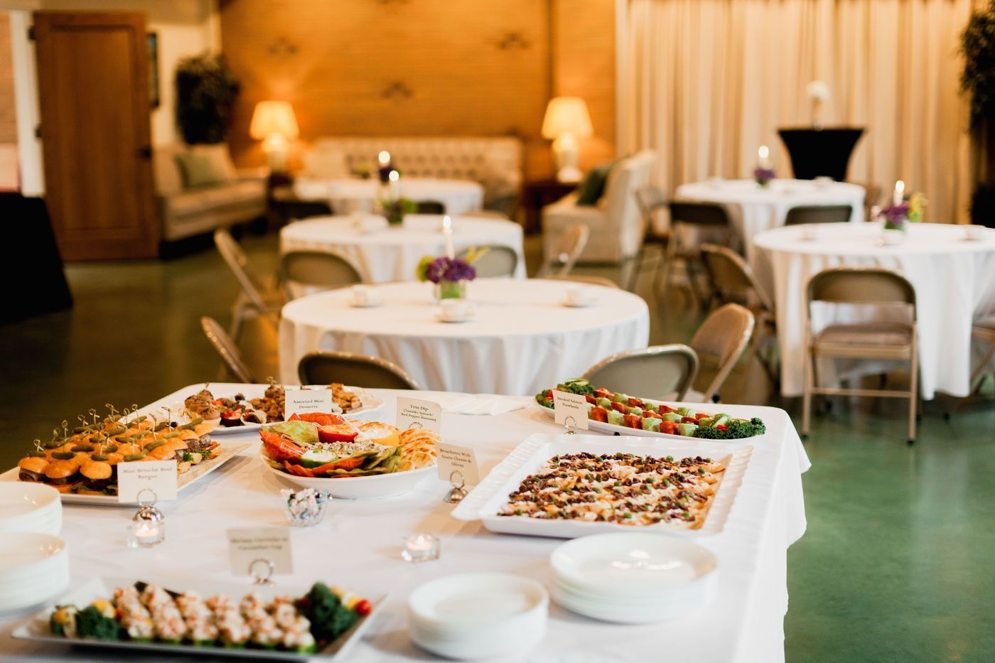 A buffet table with appetizers in the foreground, with round dining tables and seating in a softly lit event venue.