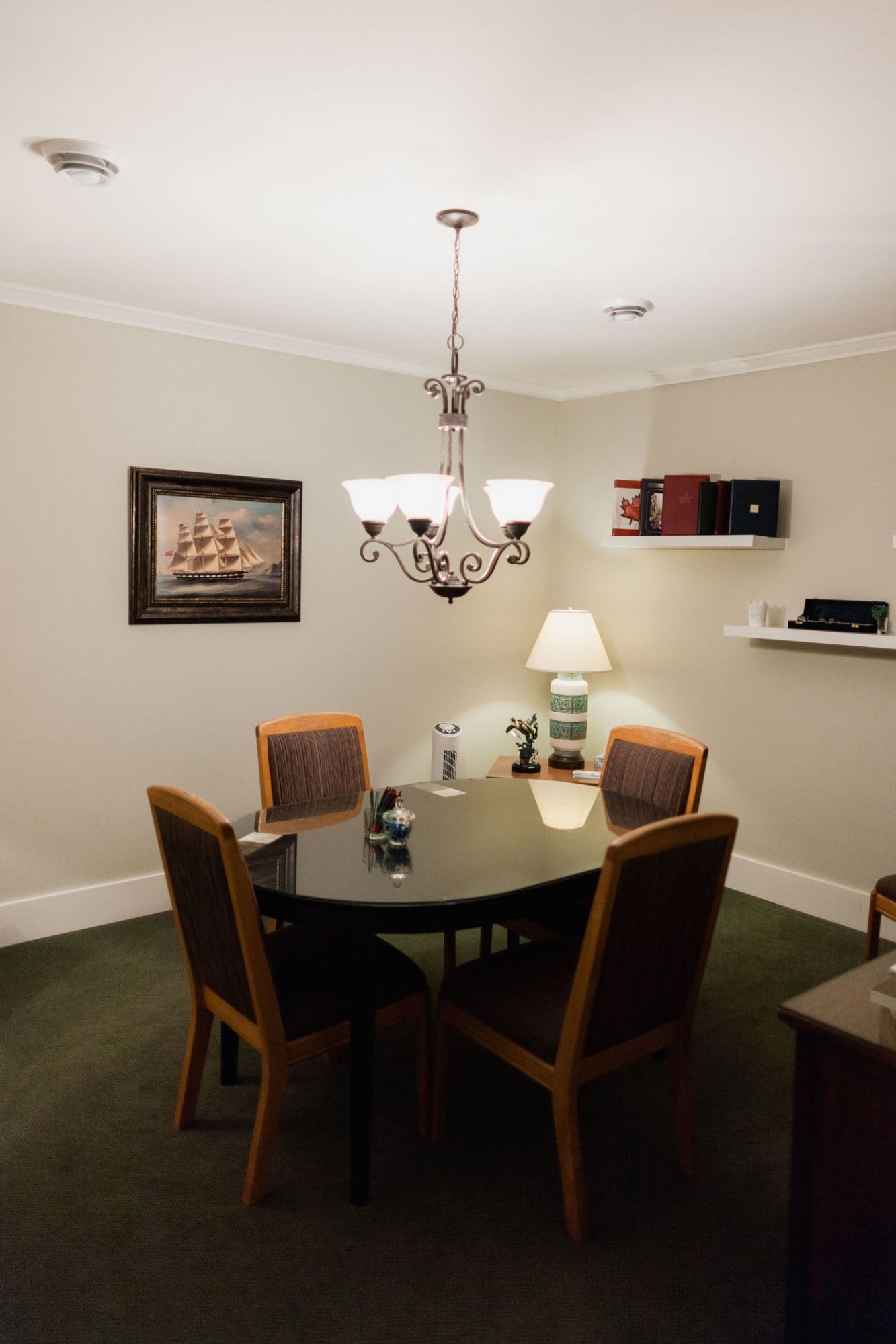 A dining area with a dark oval table, four chairs, a chandelier, a table lamp, and wall-mounted shelves on green carpet.