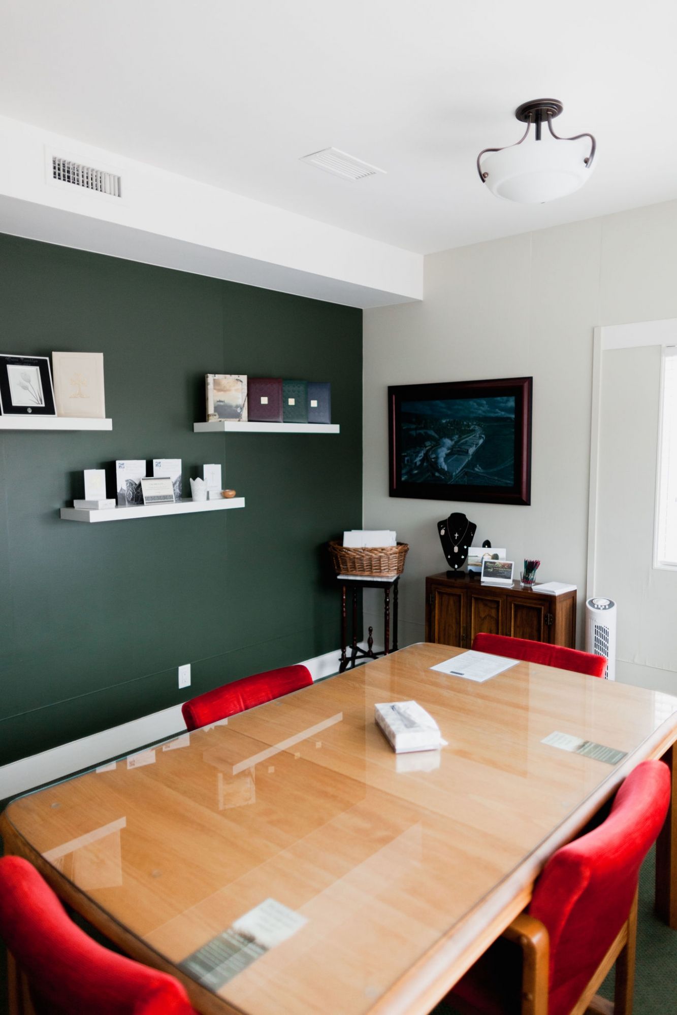 A meeting room with a dark green accent wall, white shelves, a long wooden table, and red chairs.