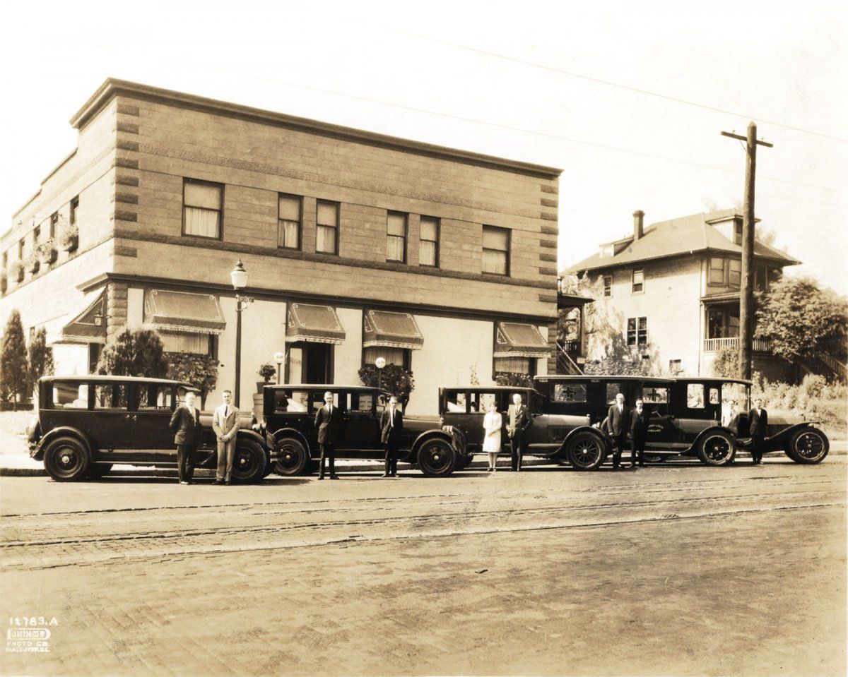 Sepia-toned photo of five vintage cars parked in a row before a brick building, with several figures standing beside them.