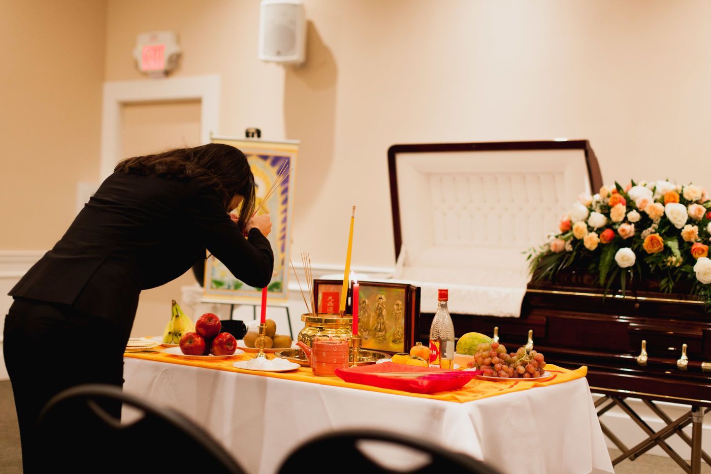 A person in dark clothing bows before an altar with candles, fruit, and offerings set up in front of an open casket.