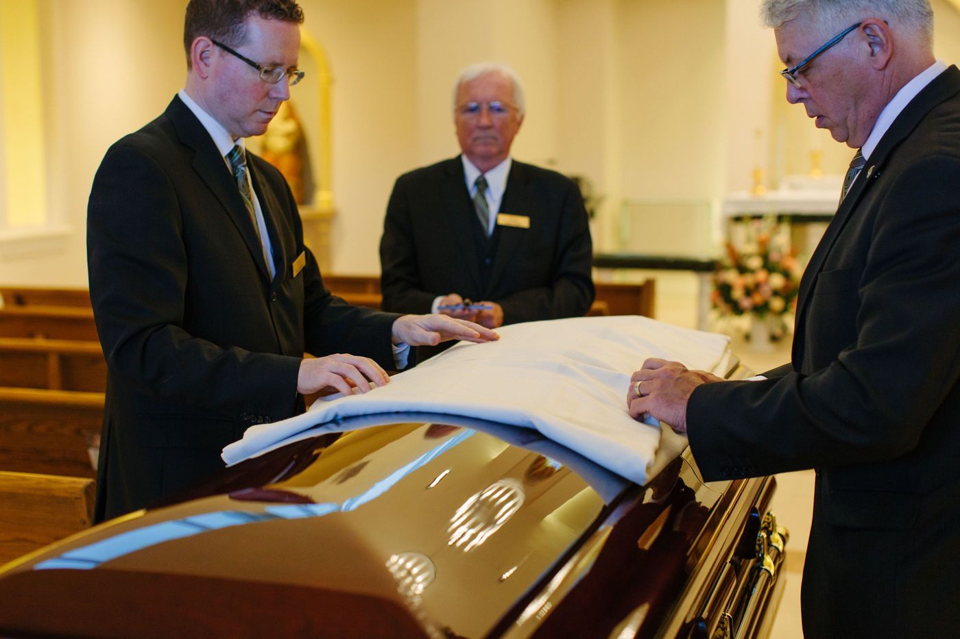 Three people in dark suits drape a white pall over a polished wooden casket in a church.