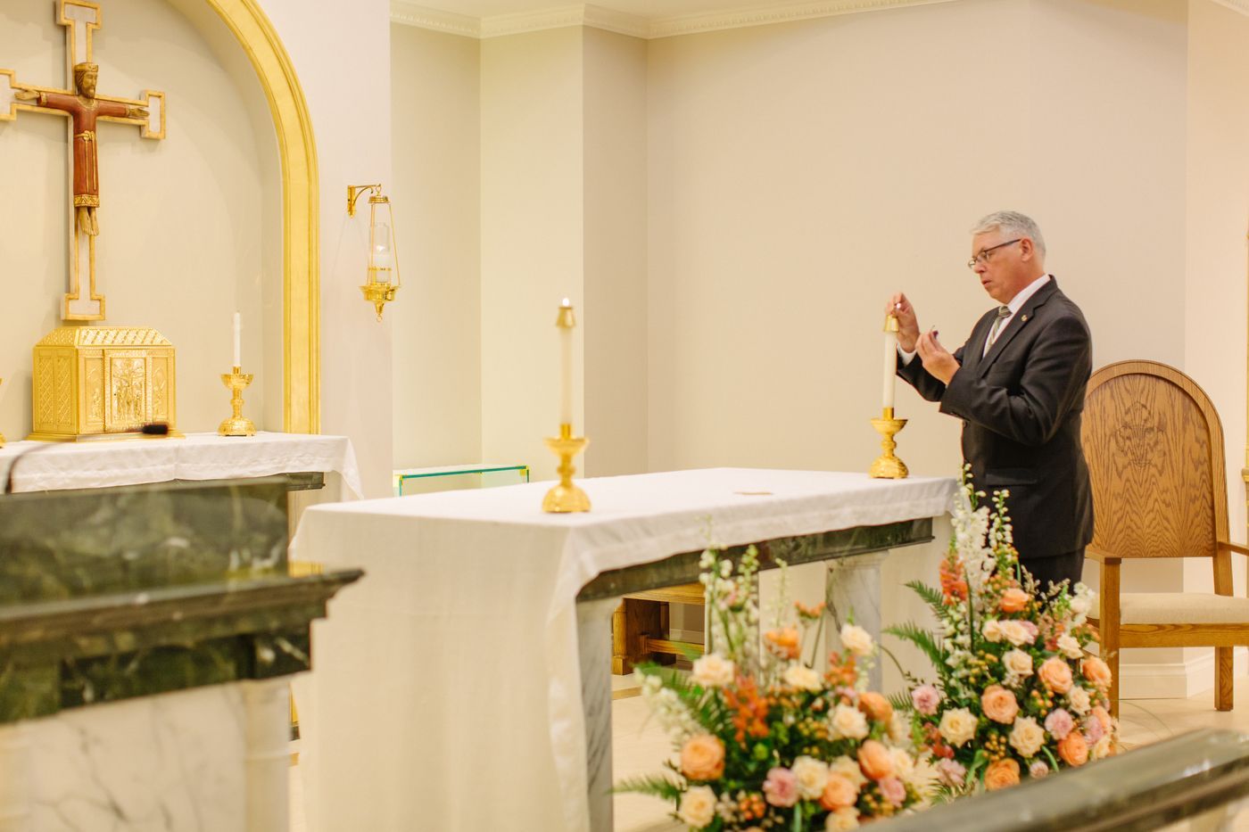 A person in a suit stands by an altar in a church, gesturing near a lit candle with floral arrangements in front.