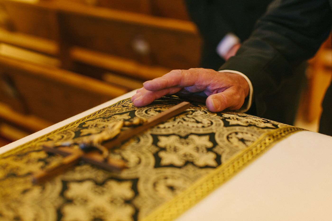 A person's hand rests on a crucifix placed over an ornate, patterned cloth, likely during a religious or funeral service.