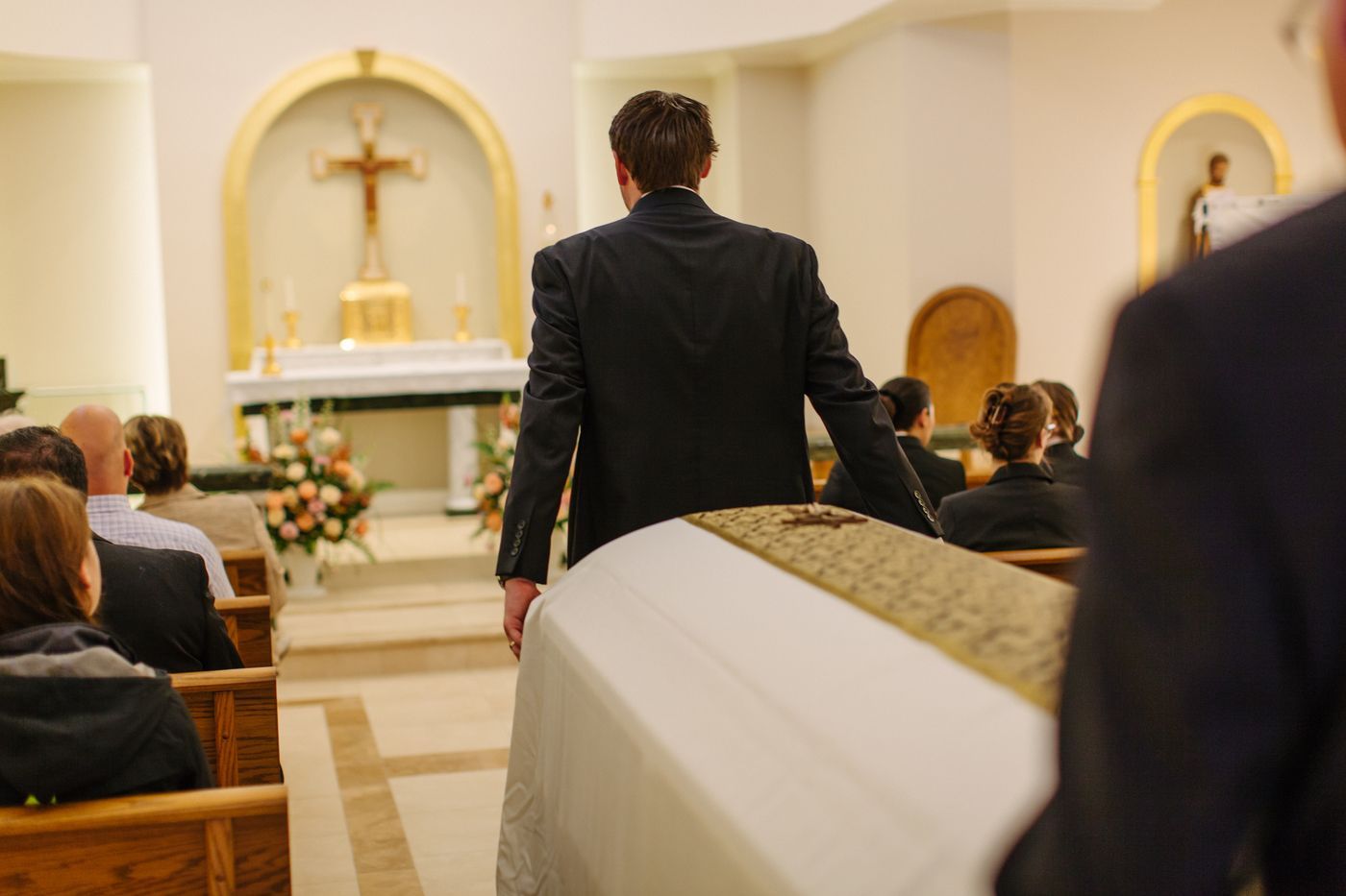 A person in a dark suit touches a casket inside a church, with attendees seated in the pews facing an altar.